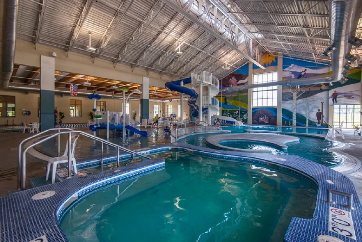 This bright, energetic indoor pool scene features children splashing down waterslides, a colorful mural in the background, and families enjoying time together in the water. The environment is lively and inviting, perfectly capturing communal recreation and the fun of the Grand Park Rec Center.