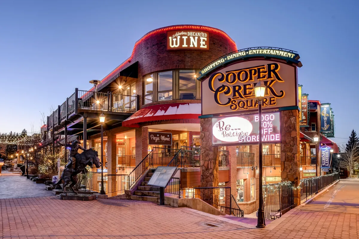 A lively exterior shot of Winter Park's Cooper Creek Square, featuring the distinctive sign, festive lights, a wine shop, and a sculpture of a horse. The atmosphere is vibrant, suggesting nearby shopping, dining, and entertainment options just moments from the villa.