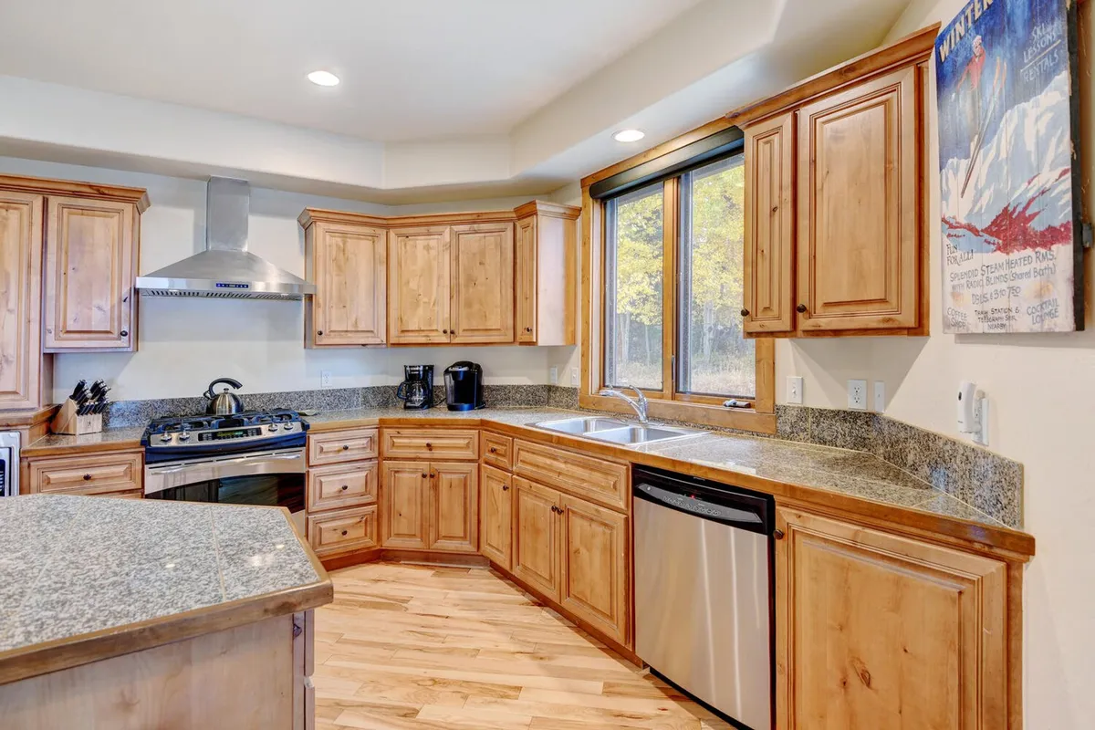 A warmly lit kitchen with granite countertops, wood cabinetry, stainless steel appliances, and a large window. Decorative wall art adds character, while the generous counter space suggests an environment ideal for meal preparation, casual family brunches, or hosting holiday feasts.