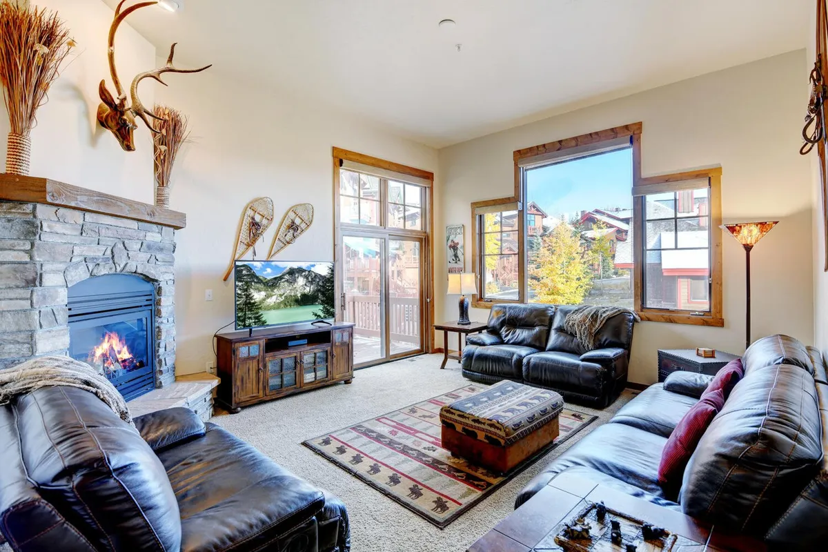 A cozy living room with a prominent gas fireplace, large leather sofas arranged around a coffee table, a patterned rug, and expansive windows revealing scenic mountain views. The space is bathed in warm light, highlighting the contemporary mountain decor and inviting atmosphere for group gatherings.