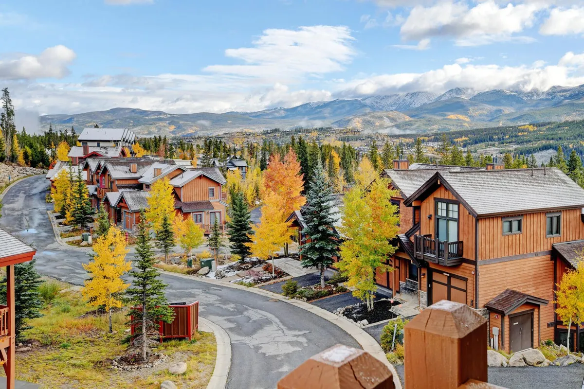 A scenic mountain village setting with colorful autumn trees, charming wooden houses, a winding road, and distant mountain peaks. The environment feels inviting for walking, biking, or simply soaking in the beauty of the Rockies.