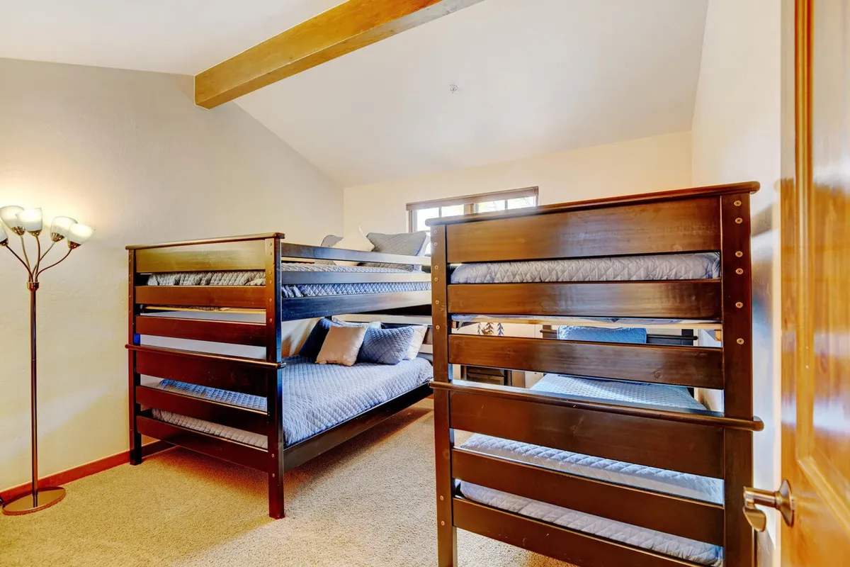 A family-friendly bunk room featuring twin-over-full bunk beds, cozy decorative pillows, and a floor lamp creating a welcoming nook. Sunlight streams through the window, illuminating comfortable spaces ideal for play, reading, or sleepovers. The layout invites both relaxation and fun for kids or families.