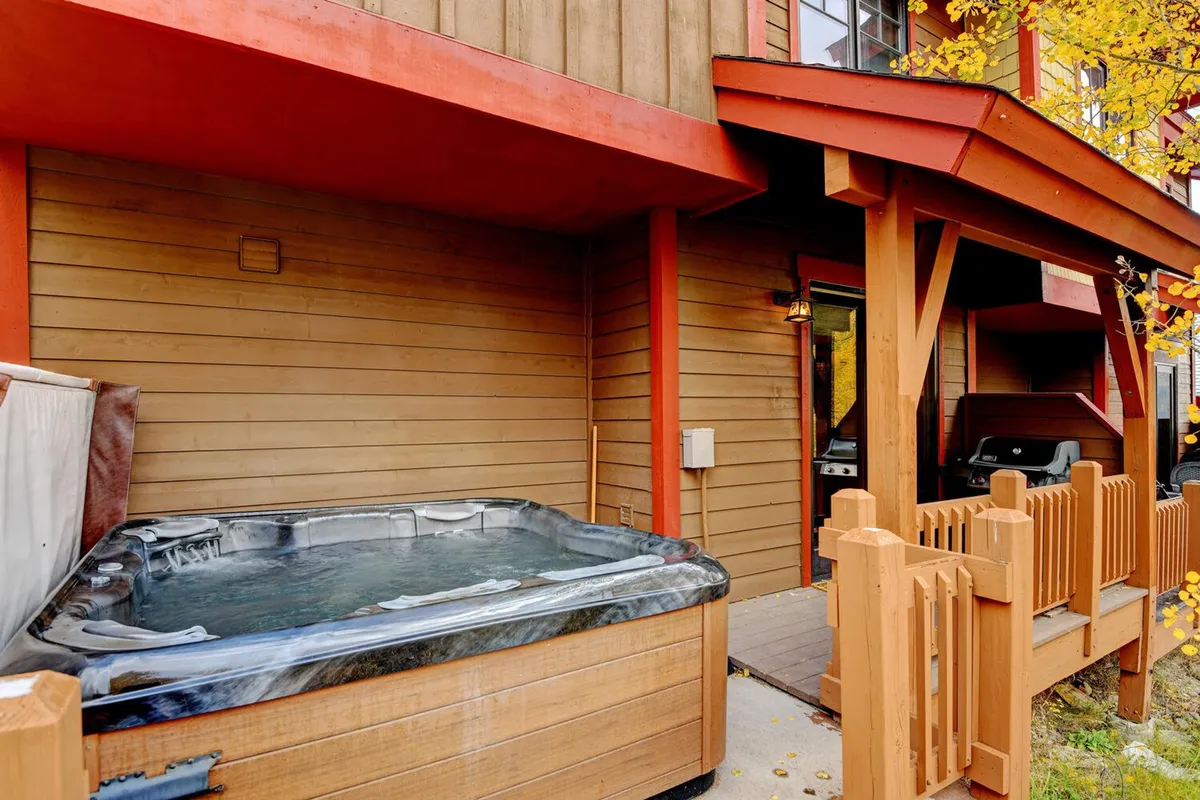 This exterior image features a cozy hot tub surrounded by wooden fencing and red accents on the building. The space is inviting for relaxing after a day of outdoor activities and is complemented by a grill set up for summer cookouts. The area hints at year-round enjoyment, with potential for stargazing on clear nights or warming up after winter adventures.