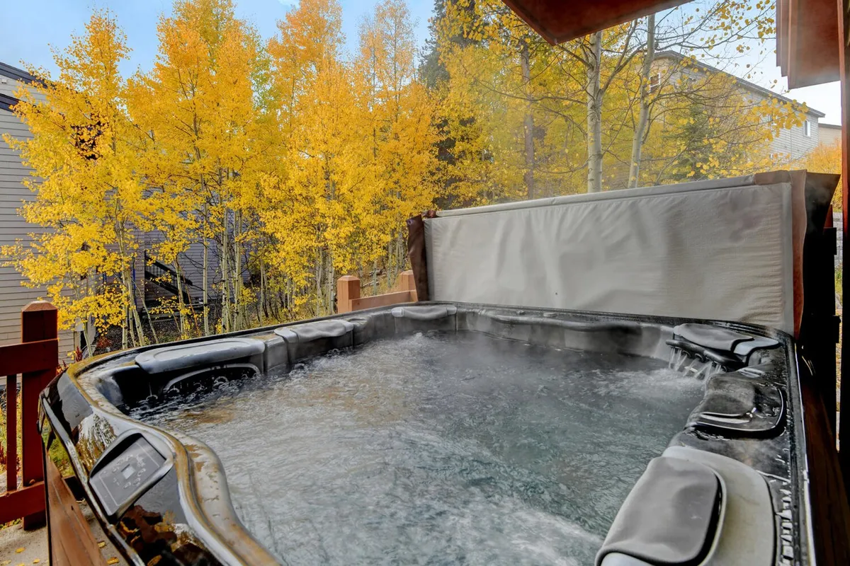 This image features a steaming hot tub set on a wooden deck surrounded by vibrant autumn foliage. The scene is serene, with inviting water reflecting the colors of the season, offering a sense of luxury and relaxation in a natural mountain setting.