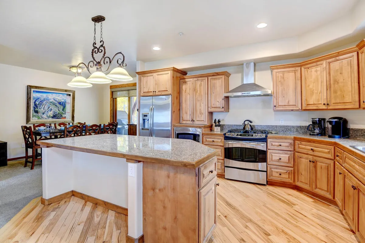This interior image features an inviting kitchen scene with a central kitchen island, warm wooden cabinets, stainless steel appliances, and a visible dining area in the background. The space is clearly designed for both meal preparation and social gatherings, offering modern amenities and an open layout that keeps the kitchen connected to the dining area.